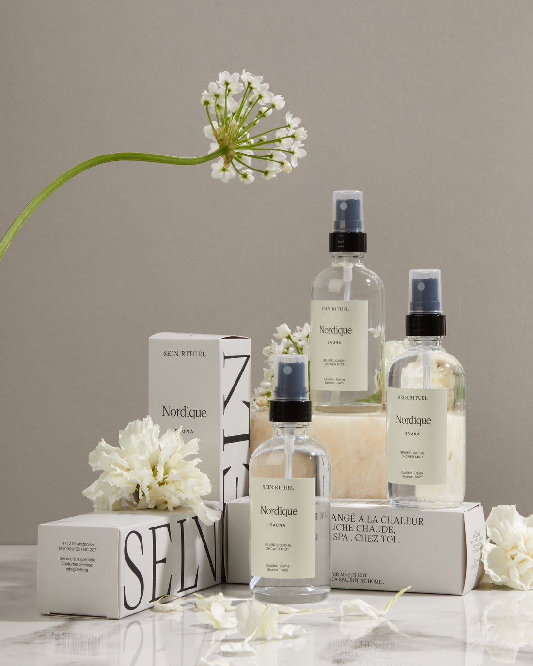 Bruines de douche Nordique Selv Rituel en flacons de verre, leurs boîtes blanches et des fleurs blanches sur un fond gris