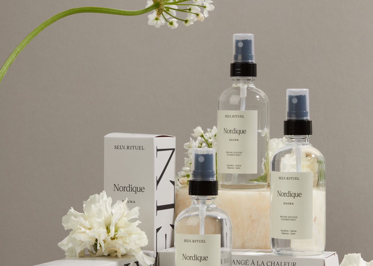 Bruines de douche Nordique Selv Rituel en flacons de verre, leurs boîtes blanches et des fleurs blanches sur un fond gris