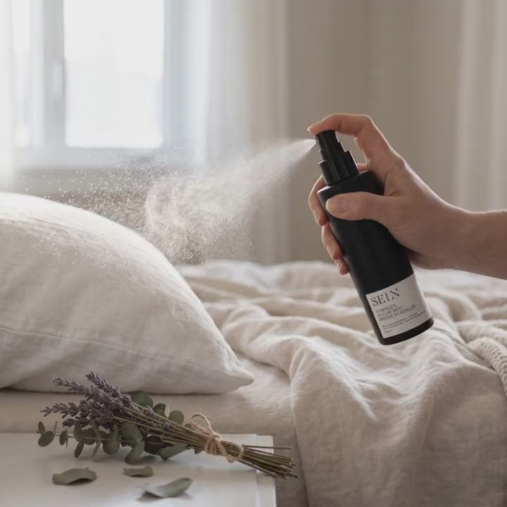 Main qui vaporise la Bruine d'oreiller Nordique Selv Rituel au-dessus d'un oreiller de lin, fine brume dans la lumière de la fenêtre, lavande et eucalyptus sur la table de chevet