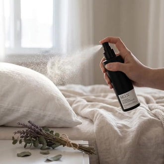 Main qui vaporise la Bruine d'oreiller Nordique Selv Rituel au-dessus d'un oreiller de lin, fine brume dans la lumière de la fenêtre, lavande et eucalyptus sur la table de chevet