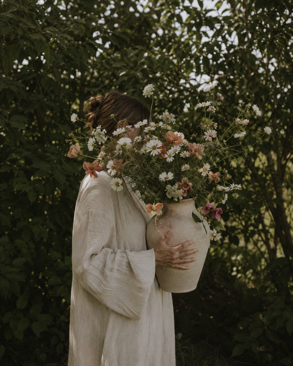 Personne en robe de lin crème tenant un grand vase de grès rempli de fleurs des champs devant un feuillage vert, visage caché par le bouquet